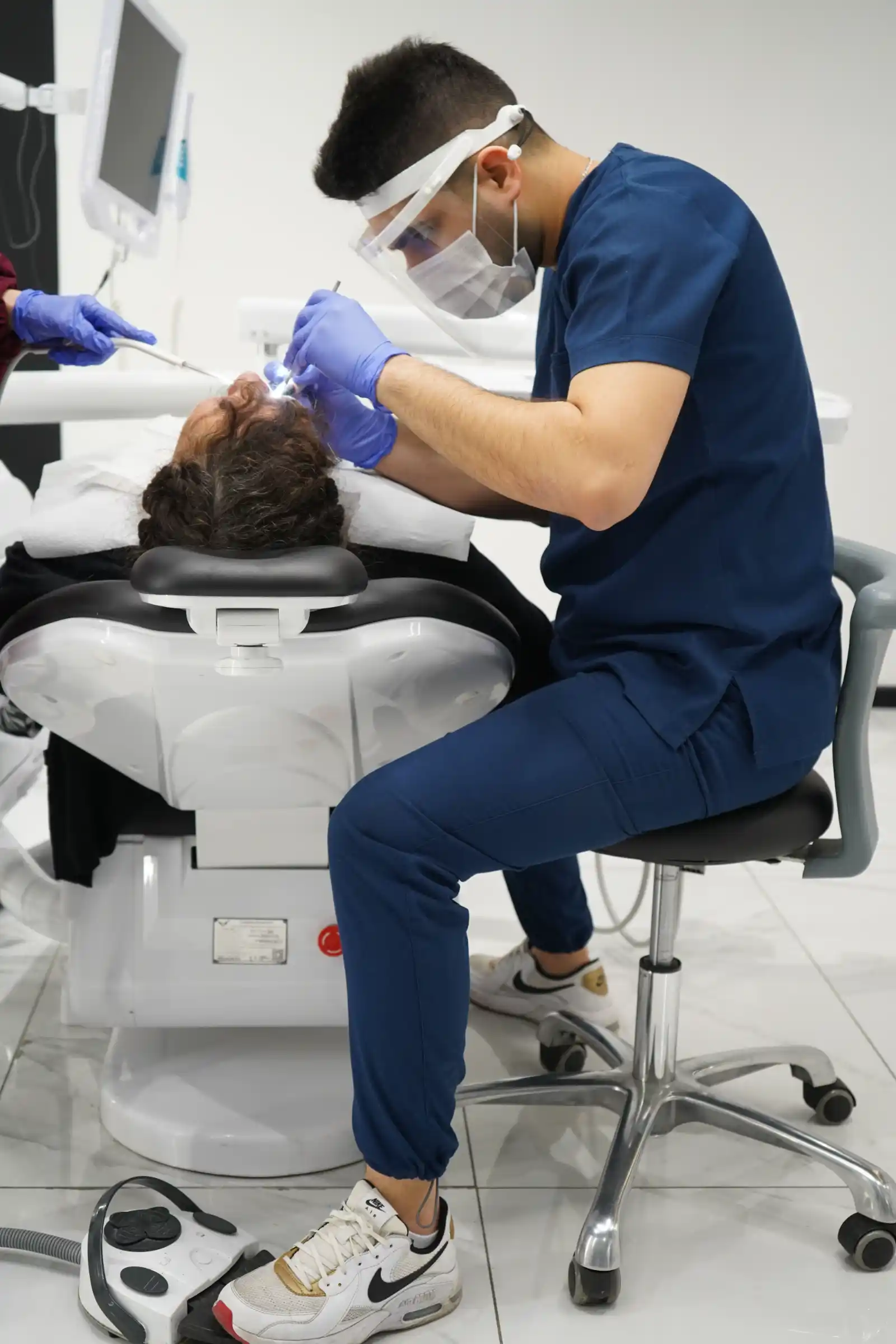 A dentist wearing a face shield and gloves treating a patient in the chair