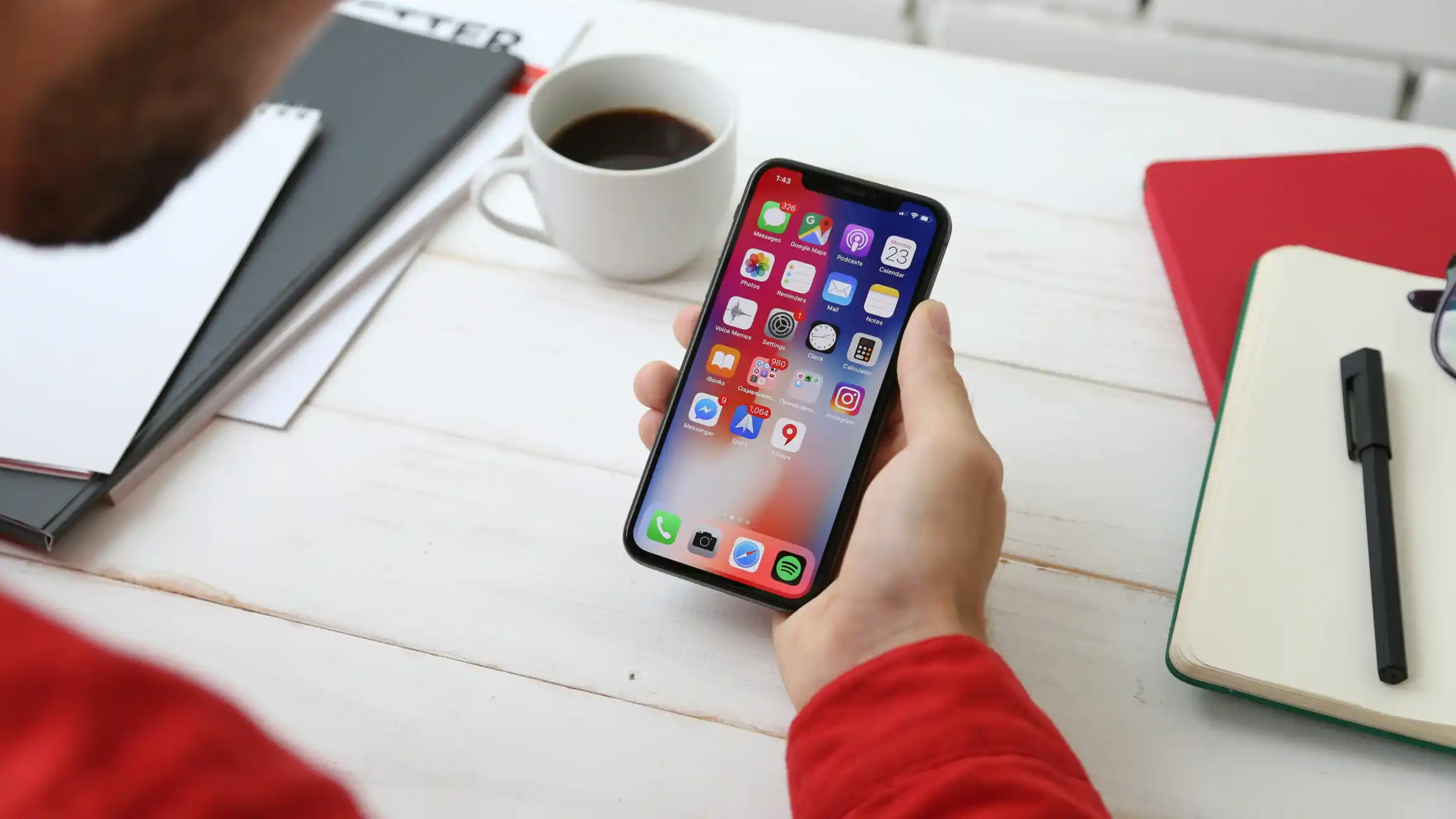A hand holding an iPhone over a desk with a notebook and a cup of coffee
